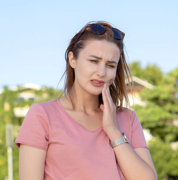 Woman in pink shirt with sunglasses on head, touching face outdoors with blurred green background.