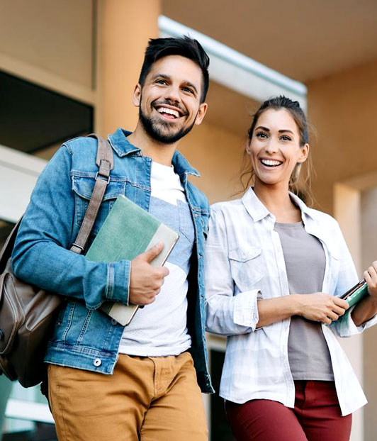 Two smiling students holding books and tablets walk together down a school hallway.