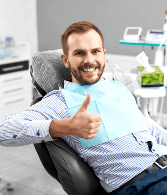 Smiling male patient giving thumbs up in modern dental office chair wearing blue shirt