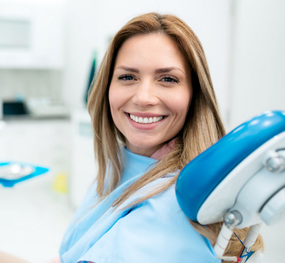 Smiling female dentist in blue scrubs holding dental tools in modern clinic setting.
