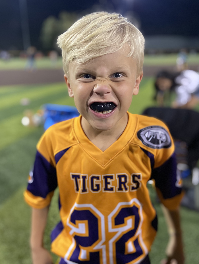 Young child wearing yellow and purple Tigers sports jersey smiling at outdoor field.