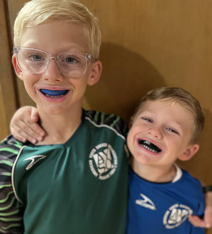 Two young boys wearing blue mouth guards smile together indoors at dental office.