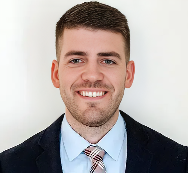 Professional headshot of smiling male dentist wearing dark blazer and light blue shirt against white background.