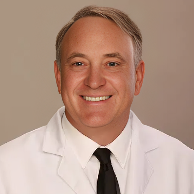 Professional headshot of smiling male dentist in white coat and black tie against neutral background.